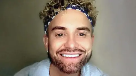 A smiling selfie of Kevin Sutherland. He is wearing a light blue shirt which is open at the neck. He has dark/ blonde curly hair and is wearing a black and white bandana with stars on it.