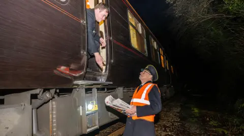 A picture of a person wearing an orange jacket delivering newspapers to the side of the Royal train.