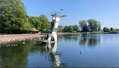 A large lake surrounded by a path and trees. It is a sunny day with birds perched on the water and a large white structure in the middle