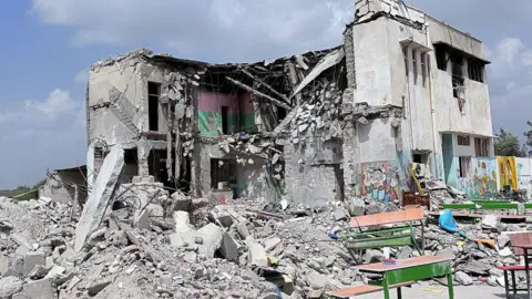A school building partially standing with walls blown off, and rubble all over the ground in front of it 