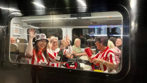 Waving Sunderland AFC fans, wearing red and white striped kit, sitting on a train. There is a group of four women - three of them wearing Sunderland colours. A woman in a hat nearest the window, on the left, holds a bottle of champagne.