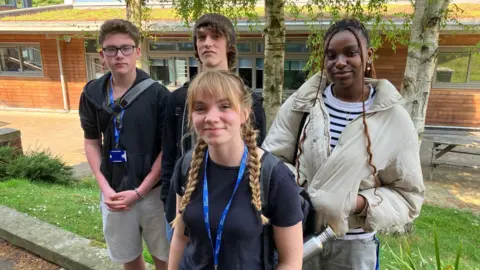 BBC A group of students stand together outside a college building. Two of them are wearing college lanyard.