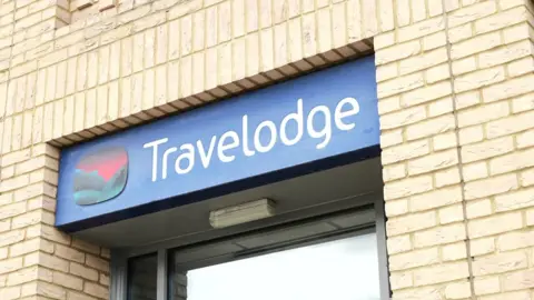 The Travelodge logo surrounded by a lightly coloured brick wall, the logo is on the entrance to a hotel