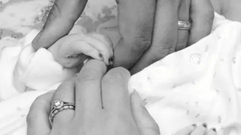 The Andrews Family A black and white photo of Wynter being held by his parents. The close-up shows their left hands holding each other and his parents' wedding rings are visible.