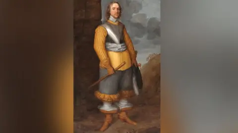 The Cromwell Museum A portrait of Oliver Cromwell, who has shoulder-length born hair, a fine lace collar done up with tassels under his neck, wearing a tawny orange/yellow doublet under a short section of body armour and a light grey silk scarf around his middle. He is wearing grey trousers which end under his knee in ruched orange material. His stockings are white and his tan-coloured boots have very wide cuffs, lined with white material. He is wearing leather gloves with fur trim and holidng a hat in his left hand and a staff in his right hand. 