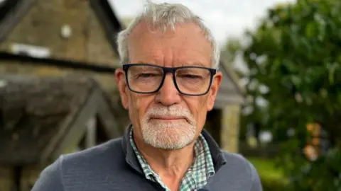 Jo Black/BBC Trevor Hicks with white hair and beard, black glasses and grey sweater. A house is visible in the background and trees to the right.