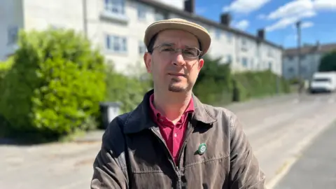 Head and shoulders image of Maurice Chiodo on Ekin Road in Cambridge. He is wearing a flat cap, thinly rounded glasses and has a goatee. He is wearing a brown jacket and red shirt.