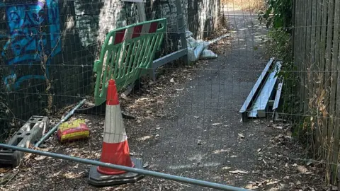 A fenced-off pedestrian path leading to the closed Oxhey Grange footbridge. A traffic cone, temporary barriers and a closure notice are visible, with trees and overgrown vegetation surrounding the route.