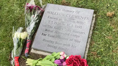 The commemorative plaque with flowers placed on it. It is square and black and says "In memory of William Clements known as Barton Billy. 7.7.1920 - 17.3.1995. 3rd the Kings own Hussars, Royal Armoured Corps. Far east prisoner of war, resting here with his loving family"