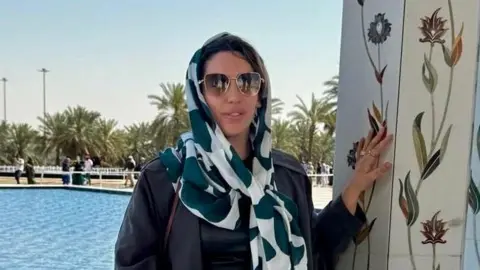 Katie Evans posa junto a una piscina y un pilar decorado con flores. Lleva un pañuelo verde y blanco sobre la cabeza, gafas de sol y un traje oscuro.