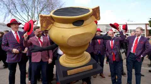 Joe Giddens/PA A group of men in red and blue checkered blazers with bright red hats. In the centre of the photo is a person dressed in a gold cup mascot suit. The men are holding their hats up and cheering with the mascot.