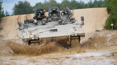 PA Media Soldiers in helmets and goggles riding in a khaki Ajax armoured vehicle. Mud is flicking up from the wet ground around the vehicle as it is in motion.