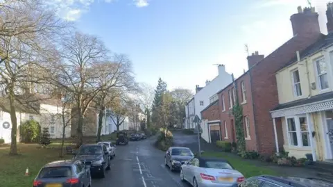 Google High Street in Shincliffe. It is a narrow two-lane road, with cars parked on each side. The street is lined with trees and houses on both sides.