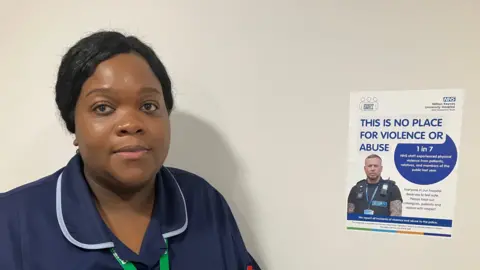 Amy Holmes/BBC A female hospital staff member in a navy uniform stands beside a wall displaying an NHS poster that promotes zero tolerance for violence and abuse. The staff member’s badge and lanyard are visible, and the poster highlights safety messages for colleagues, patients, and visitors.