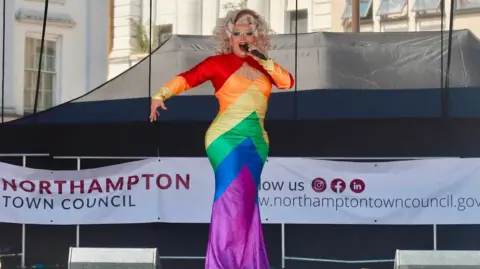 West Northamptonshire Council A drag queen dressed in a multi-coloured dress with one arm out and one hand holding a microphone, performing on a stage. A Northampton Town Council banner is at the back of the stage.