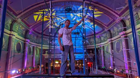 A man wearing a white t-shirt and cargo pants stands on top of trapeze equipment inside the Empire building in Great Yarmouth, with colourful lighting and light projections visible inside the ornate Victorian venue