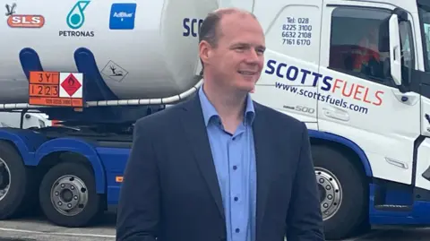 Communities Minister Gordon Lyons, wearing a blue shirt and navy suit, standing in front of a white truck. 