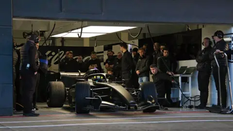 CADILLAC FORMULA 1 TEAM The Cadillac F1 car in a garage surrounded by the team. The car is black and the team surrounding it are mainly wearing black clothing.