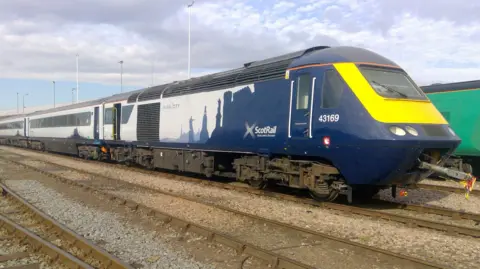 ScotRail A ScotRail Inter7City train in blue and white livery is parked on railway sidings, with multiple carriages visible along the track under a partly cloudy sky.