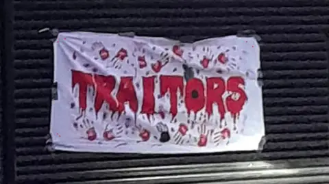 A banner stuck to black shutters. It reads "Traitors" in red writing made to look like blood and with bloody hand prints. The "O" is styled as a Remembrance poppy.