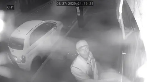 Metropolitan Police Black and white image of Muzyka looking up into the camera at a door. He is wearing a baseball cap and light-coloured clothes. A small parked car is also visible behind him.