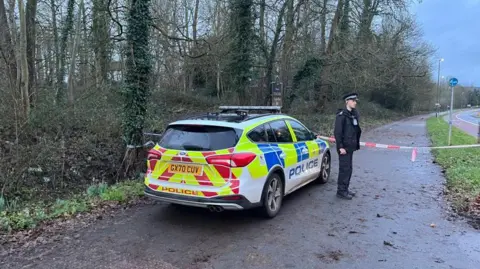 A police officer stands next to a patrol car, with a taped off area behind him. 