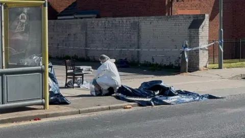 Jonny Humphries/BBC A forensic investigator in white PPE collects evidence on the kerbside next to a yellow bus-stop. A wooden chair used to help the victims and a blue tarpaulin is lying nearby