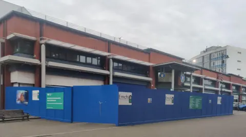 The entrance to Barrow Market Hall. The long two-storey redbrick building has a sign above the doorway and is surrounded by blue wooden boards. Street lights can be seen on the flat roof.