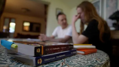 A father and son sat at a kitchen table, with one resting their forehead on their hand, in soft focus. Two GCSE text books are in the foreground.