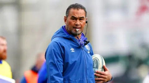 Pat Lam, wearing a blue coat and holding a rugby ball. There is a whistle in his mouth.