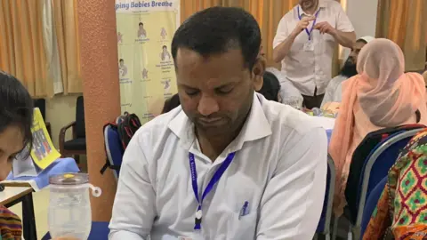 Michael Fernando A woman at a table, next to a man and another woman. She is using breathing apparatus on a baby medical doll