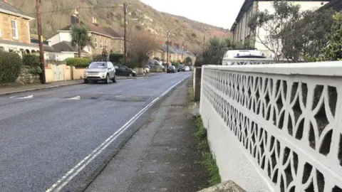 Penberthy Road in Portreath. It is lined with houses and cars parked along it. A hill can be seen in the background.
