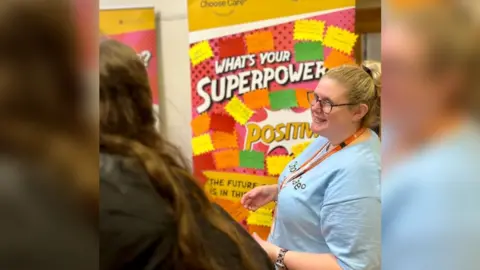 Two women, one with brown hair wearing a black coat with her back facing the screen. Another with blonde hair wearing a blue t-shirt, an orange lanyard and glasses. A colourful board in the background reading 'What's your superpower'.