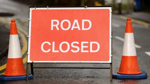 Road closed sign. The red and white sign has been placed in the middle of a road. Two traffic cones have been placed either side of the sign.
