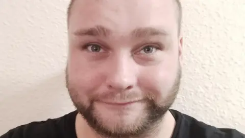 A close-up selfie of a man with a light brown beard and light-brown eyes. He is smiling at the camera. There is a white wall behind him.