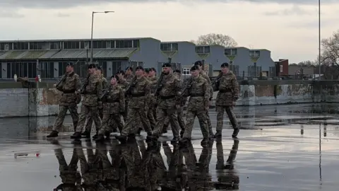 A group of soldier, wearing camouflage, with a rifle on their right shoulder marching during a training operation. They are marching outside on a dark tarmac surface. There are five buildings in the background that are either two or three storeys. 