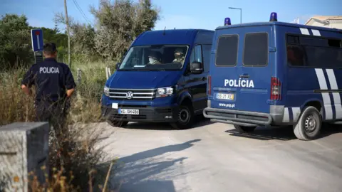 Reuters Two vans with German license plates arrive at a blocked road. They are blue with white writing. A male policeman stands with his back to the camera on the left of the image