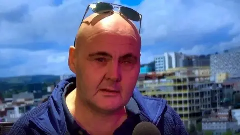 Mr Roberts sits in front of a microphone with a picture of Sheffield's skyline spread across the wall in the background. He is bald and has sunglasses pushed back over his head.