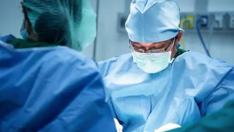 A surgeon wearing glasses and blue scrubs looks down at his hands as he operates on a patient