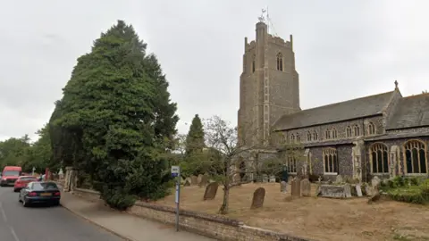 Google A general view of Church Street in Stradbroke. To the right is the large, church, made out of grey and brown stones, and its spire. There are various graves dotted throughout the cemetery in front of the church. To the left is the road, with three cars parked next to the kerb.