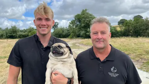 Ellen Knight/BBC Will and Rob stood side by side, both wearing black polo shirts. Will has short blond hair, and Rob has short grey hair. Will is holding a black-and-tan pug dog. They are both stood on the bank of the new meander and are smiling widely. 