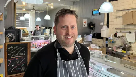 Richard Hards in a grey apron smiles at the camera. The photo is taken inside a brightly lit butcher's shop, where another man in the same uniform is stood behind a counter.