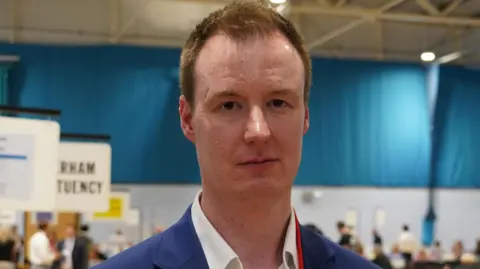 Chris Read poses for a photo, inside a sports hall. He has short ginger hair and is wearing a blue suit and white shirt. A red lanyard hangs round his neck.