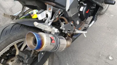 The picture shows the rear right-hand side of a black sports motorcycle parked on a road surface. The most noticeable feature is an aftermarket exhaust canister with a blue-tinted metal tip and a visible "Black Widow" logo, along with some rust and wear around parts of the frame and exhaust system.