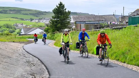 Five cyclists using the revamped cycle path in Lowca. A group of three is in the foreground, with two cyclists a short distance behind them. There are houses on the right hand side and in the distance, surrounded by lush green countryside. All the cyclists are wearing helmets and colourful jackets, some have bags attached to their bikes.
