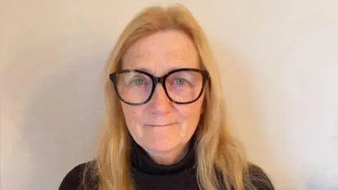 An elderly woman, blonde, with thick black glasses standing against a wall wearing a black polo neck top