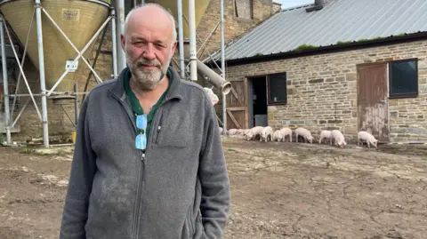 A balding man with a white beard is wearing a grey fleece with a pair of glasses hung from the zip. He is stood on a farm with a number of pigs in the background and farm buildings