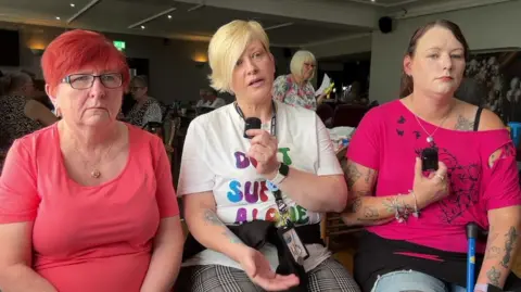 Julie, Kerrie and Vicky sit in a row together in a busy pub. They look at the camera, with Kerrie speaking in the middle. They are sitting down. Two have pink T-shirts while Kerrie has a white one with "Don't suffer alone" written on it.