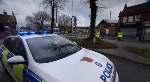 The approach to a roundabout, with two large trees visible. There is a police car with its light flashing and two officers wearing a bright yellow jacket standing in front of a row of shops