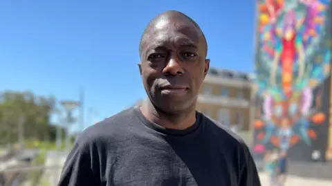 Stuart Woodward/BBC Bayo Alaba, a man wearing a black T-shirt, looks directly at the camera. Behind him, blurred, is a large mural on a wall.